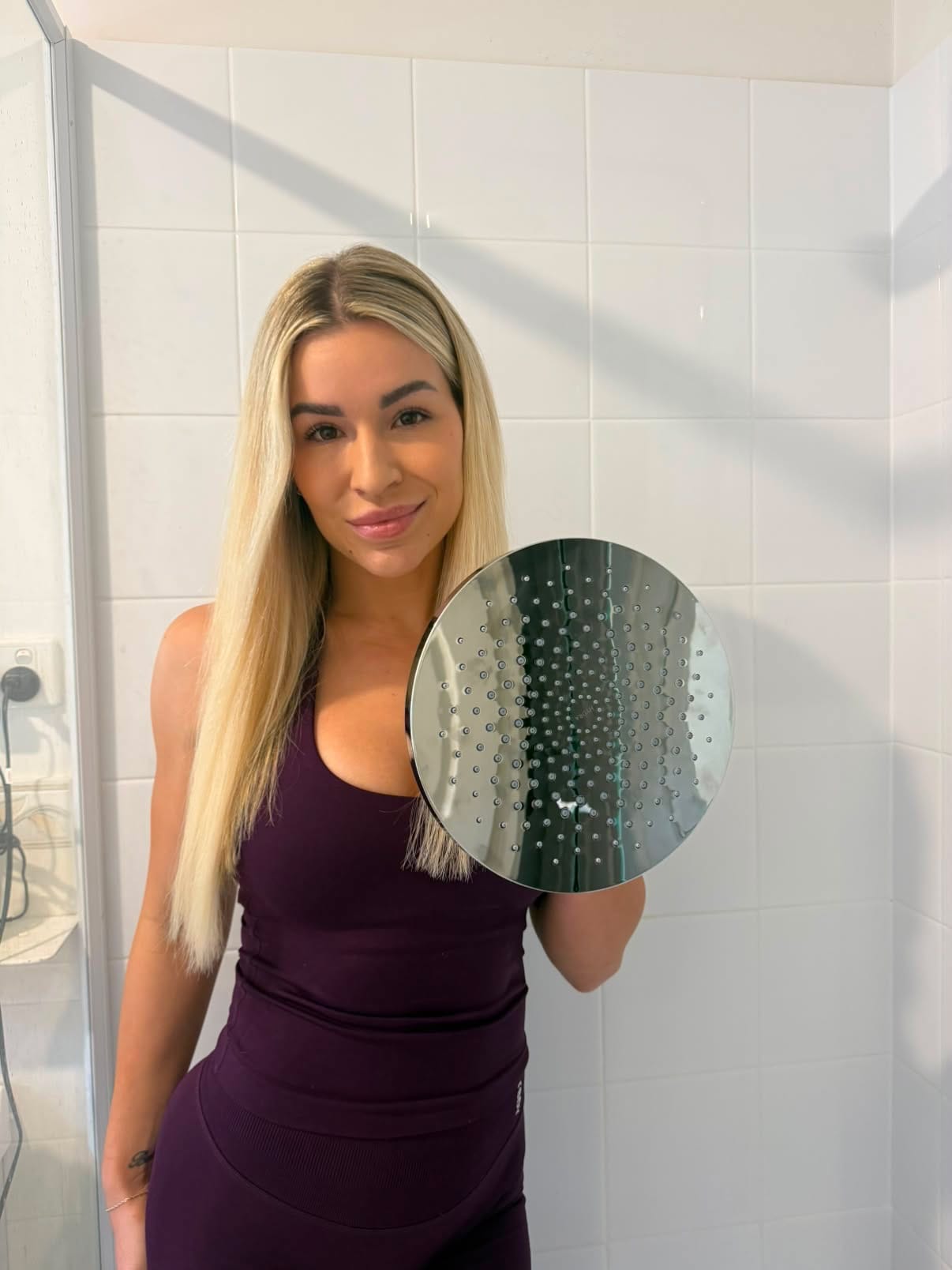 A woman with long blonde hair stands in a tiled bathroom holding a round mirror. She is wearing a dark purple sleeveless dress.